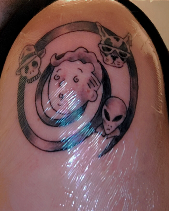 A tattoo artist's work with a cartoon face, skull, dog, and alien on a person's shoulder, a questionable tattoo idea.