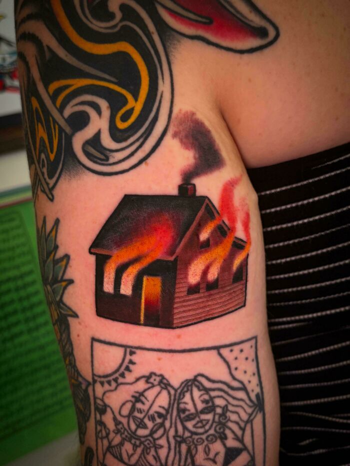 A colorful tattoo of a house on fire next to other tattoos, illustrating a tattoo artist's questionable talent.