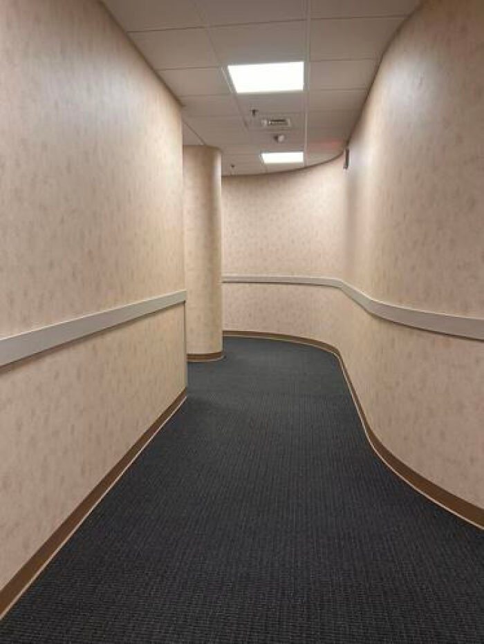 A long, curved, haunting liminal space in a hospital-like corridor with textured walls and dark carpet.