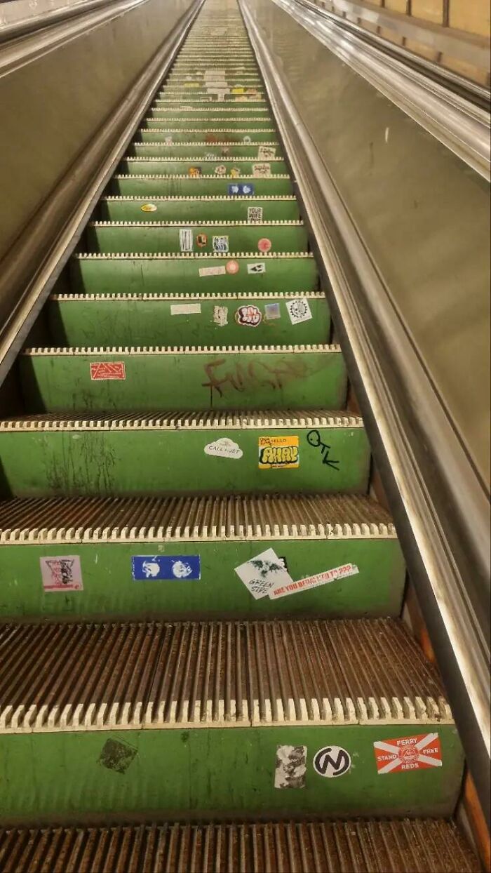 A long escalator with green steps is covered in numerous stickers and graffiti, a bizarre sight.