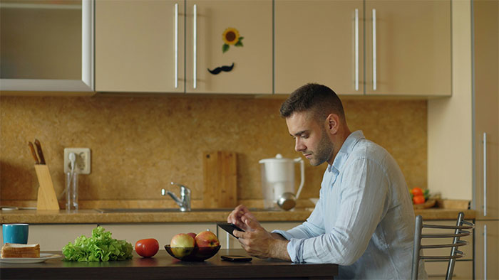 Mom's Boyfriend sits at a kitchen table, looking at his phone, while testing a kid's food issue.