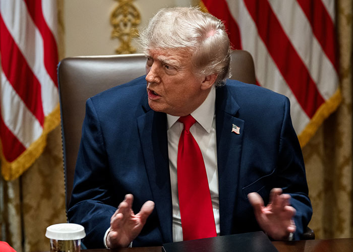 Trump discussing at a meeting with US flags in background Trump discussing at a meeting with US flags in background