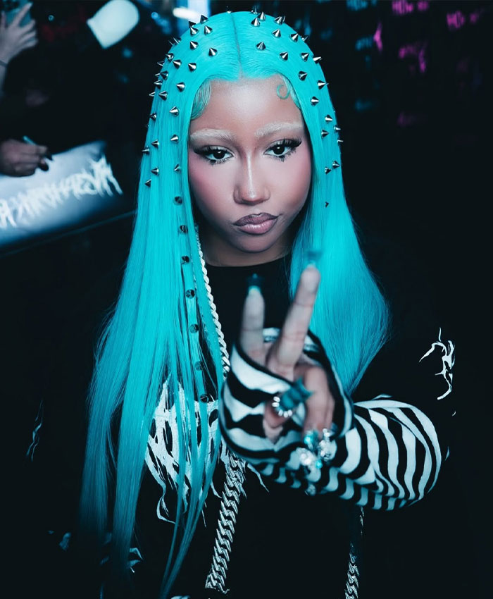 A woman with blue, spiked hair and a bold outfit, giving a peace sign.