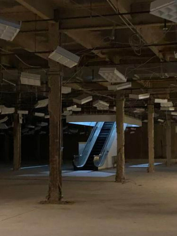 A vast, dark, abandoned space with exposed ceilings, columns, and an escalator, a haunting liminal space.