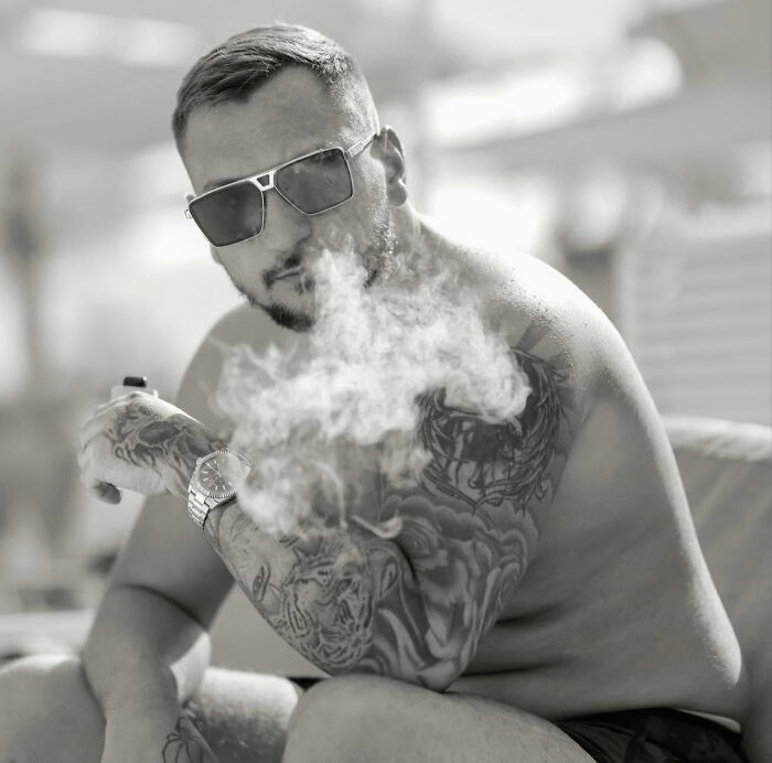 Male influencer with tattoos and sunglasses exhaling smoke, reflecting grave charges after being arrested in Dubai.