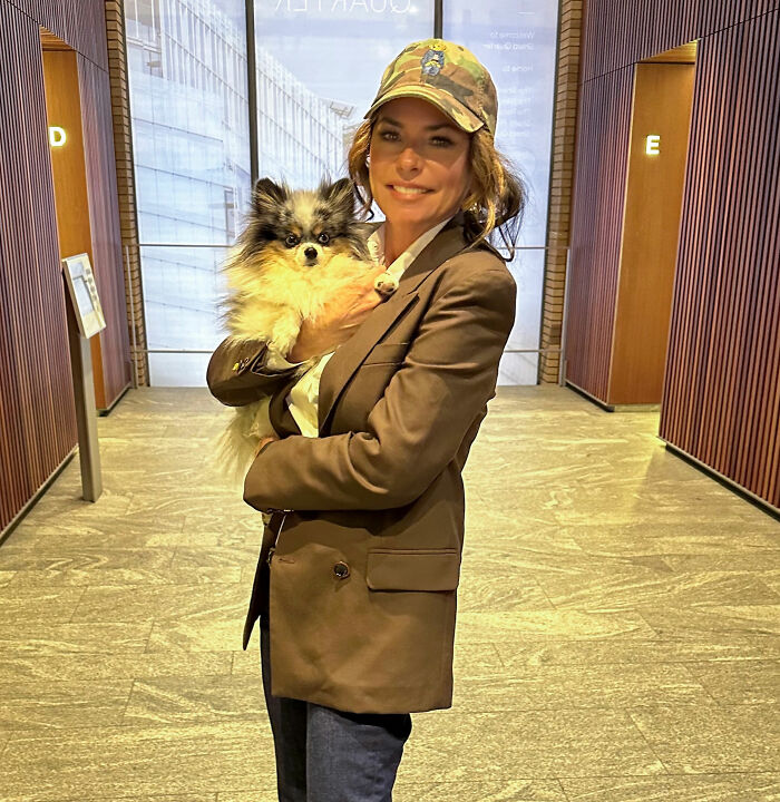 Shania Twain holding a small dog, wearing a hat and jacket, showing her transformation aftermath. Shania Twain holding a small dog, wearing a hat and jacket, showing her transformation aftermath.