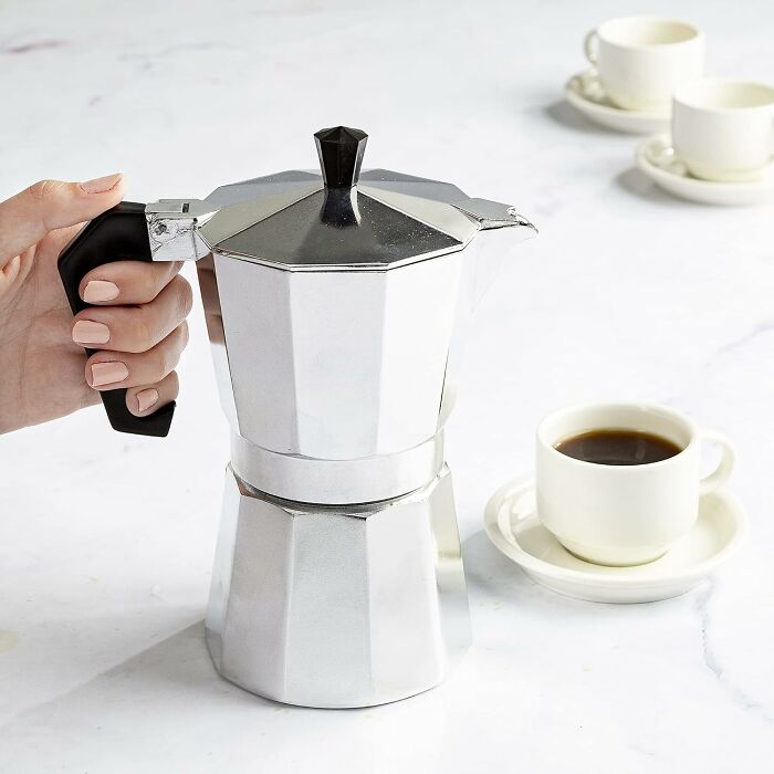 Hand holding a silver Moka pot next to a small cup of coffee, part of Prime Day Deals for coffee lovers.