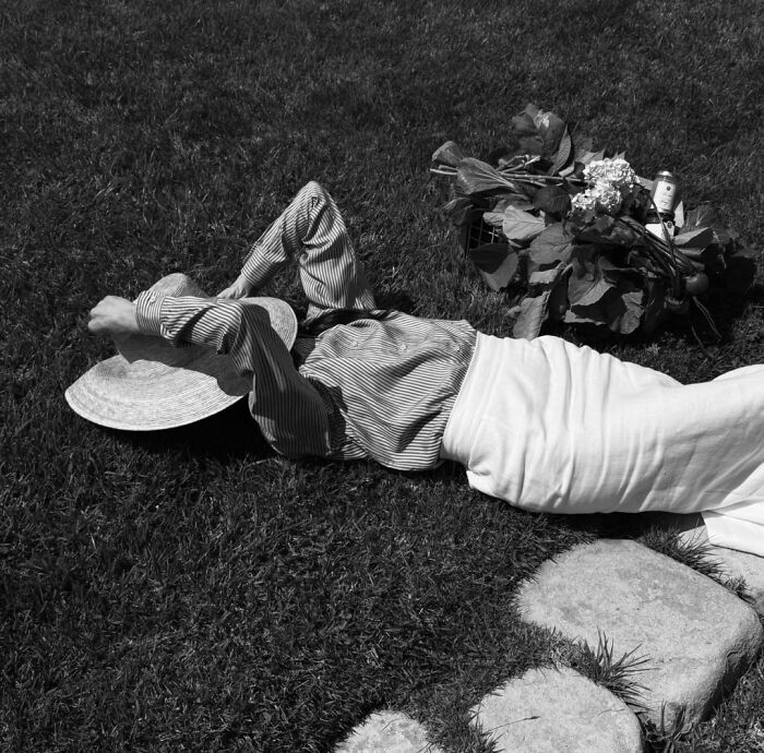 Black and white photo of a woman lying on grass with a hat and plants nearby
