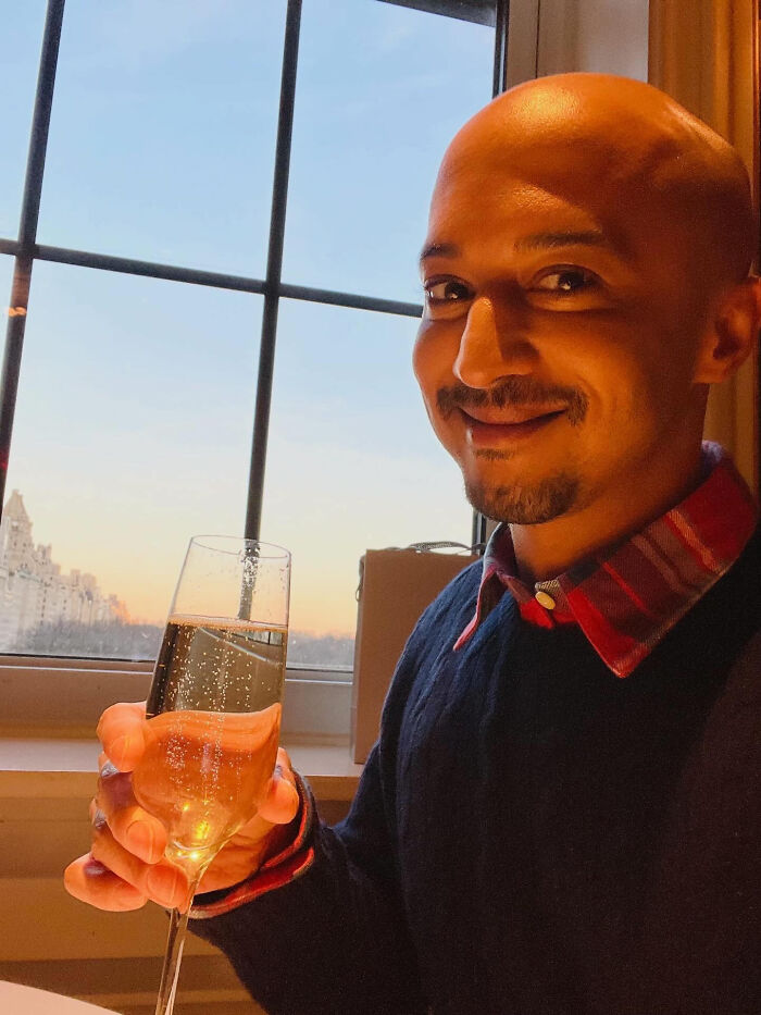 A man holding a glass of champagne, smiling at the camera, with a city view outside the window. This shows a wild coincidence.
