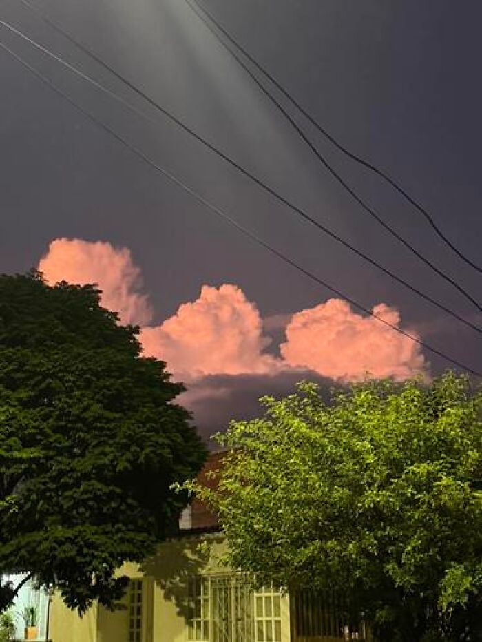 Haunting liminal spaces in the night sky with large, pink-glowing clouds over trees and a building.
