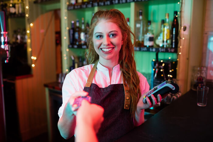 A smiling bartender with red hair processes a credit card payment, showcasing customer service and disturbing things people learned about their friends.