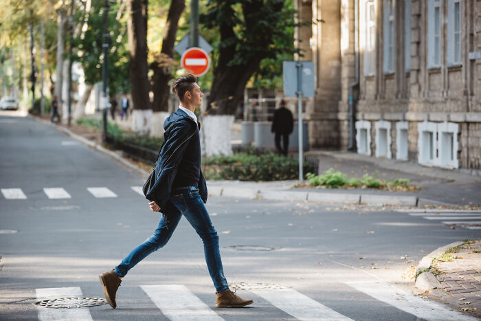 A young man in a dark jacket and jeans strides across a crosswalk on a city street, symbolizing disturbing things people learned about their friends.