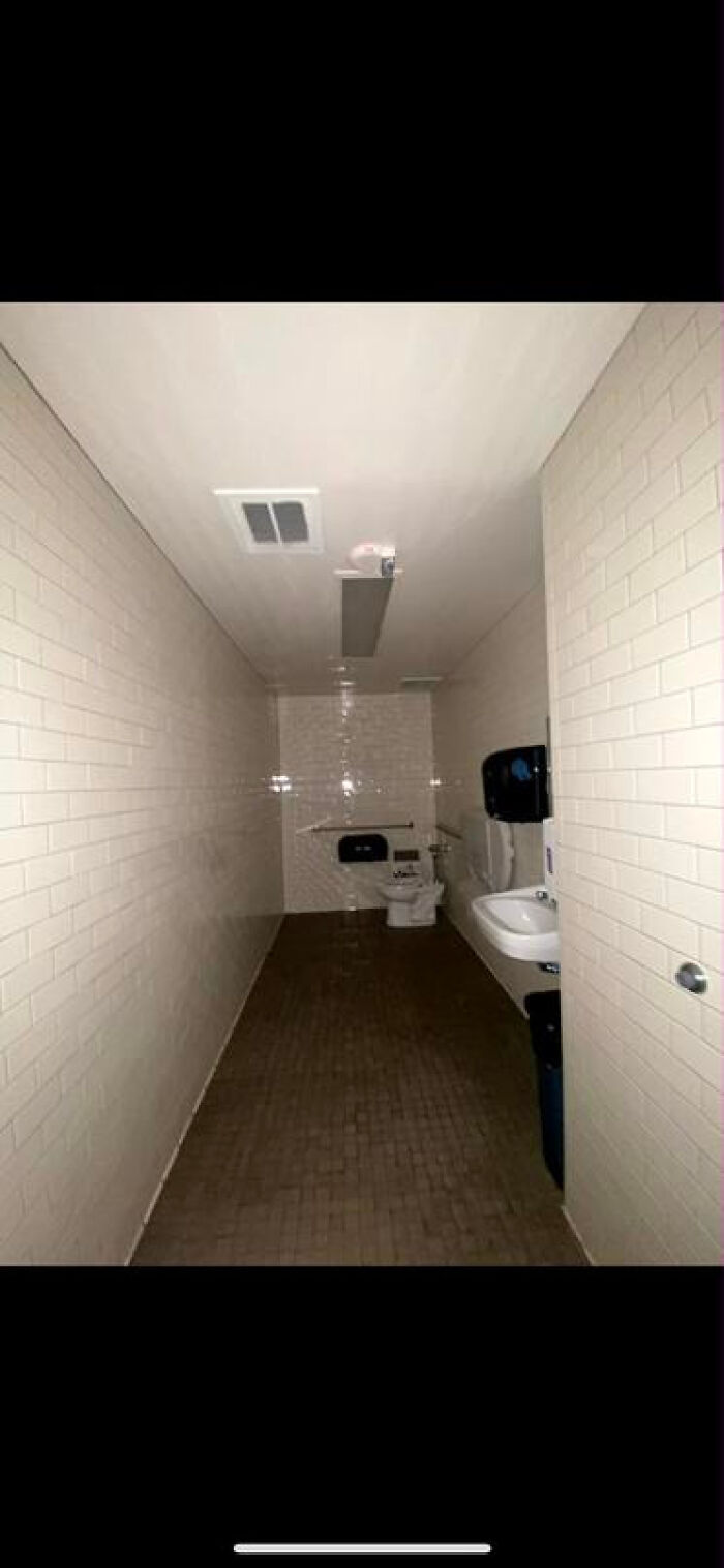 A narrow, tiled public restroom, dimly lit, evoking an unsettling liminal space.