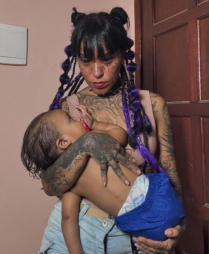 Woman with tattooed eyes holding a baby, highlighting her extensive body art.