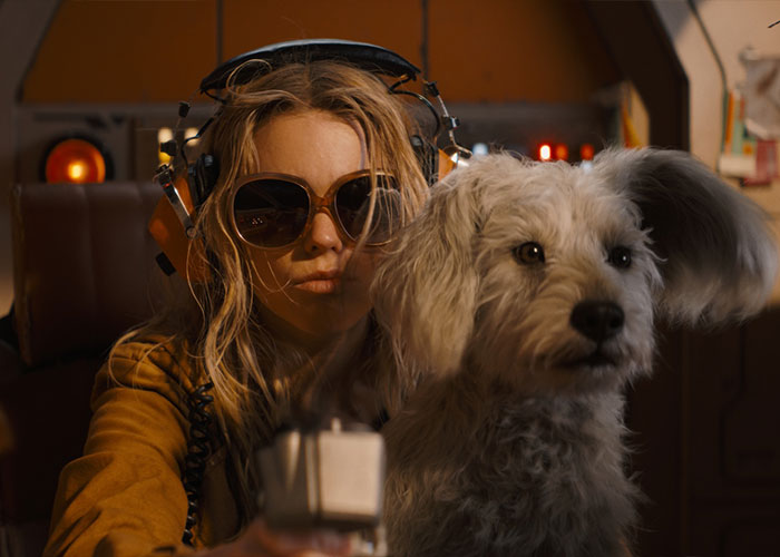 Supergirl fans watch Supergirl in oversized headphones and sunglasses, holding a small white dog.