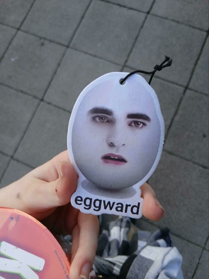A person holding a bizarre egg-shaped air freshener with a man's face and the word eggward, raising questions.