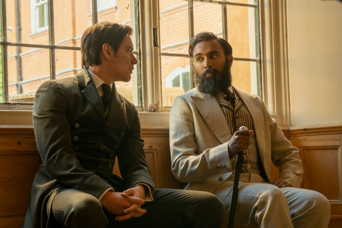 Louis Partridge and a man with a beard sit by a window, talking in a still from the Enola Holmes 3 movie. Louis Partridge and a man with a beard sit by a window, talking in a still from the Enola Holmes 3 movie.