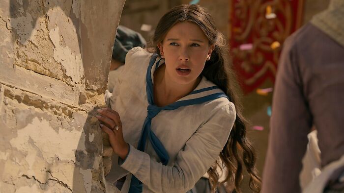 Millie Bobby Brown looks concerned, peeking around a crumbling wall in a scene from the Enola Holmes 3 movie. Millie Bobby Brown looks concerned, peeking around a crumbling wall in a scene from the Enola Holmes 3 movie.