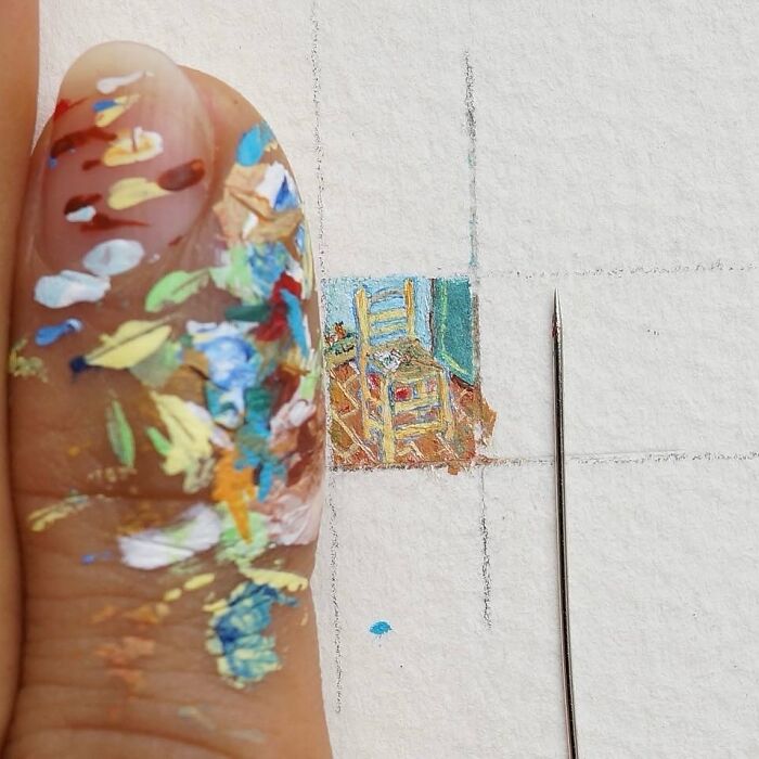 A thumb covered in various paint swatches next to a minuscule, detailed miniature paper figures painting of a chair, with a fine needle.
