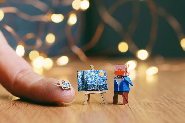 A miniature paper figures artist painting a starry night scene on a tiny easel, with a thumb holding a tiny palette.