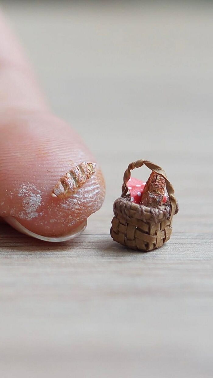 A miniature paper figures basket filled with bread and a red cloth sits next to a human fingertip.