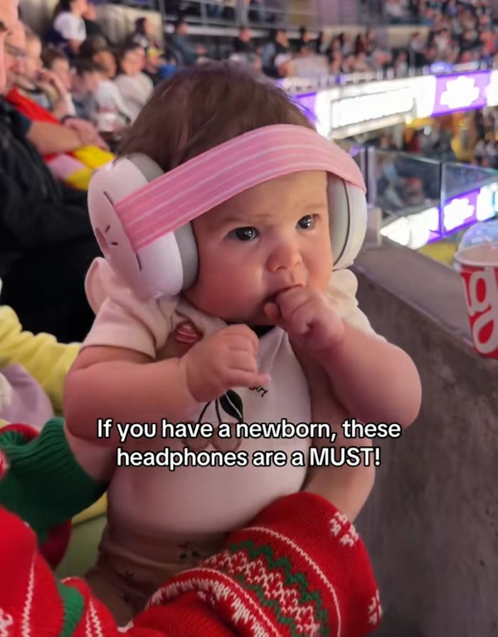 A baby with ear muffs at World Cup. The baby wears pink and white ear muffs, with a pink headband, and is held by an adult wearing festive red and green attire.