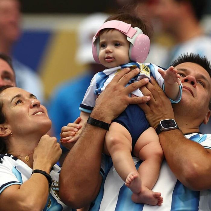 A baby wearing ear muffs at World Cup. The baby, dressed in an Argentina jersey, is held up by an adult, while a woman looks on with a smile.