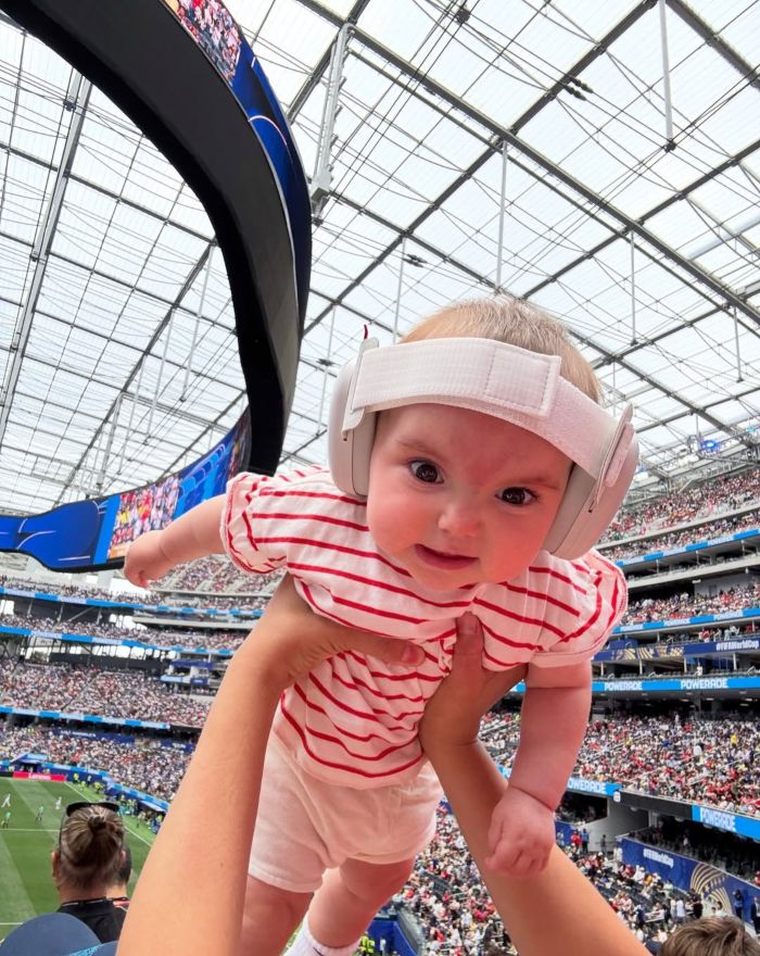 Baby with ear muffs at World Cup, held up high in a stadium, with an alert from doctors.
