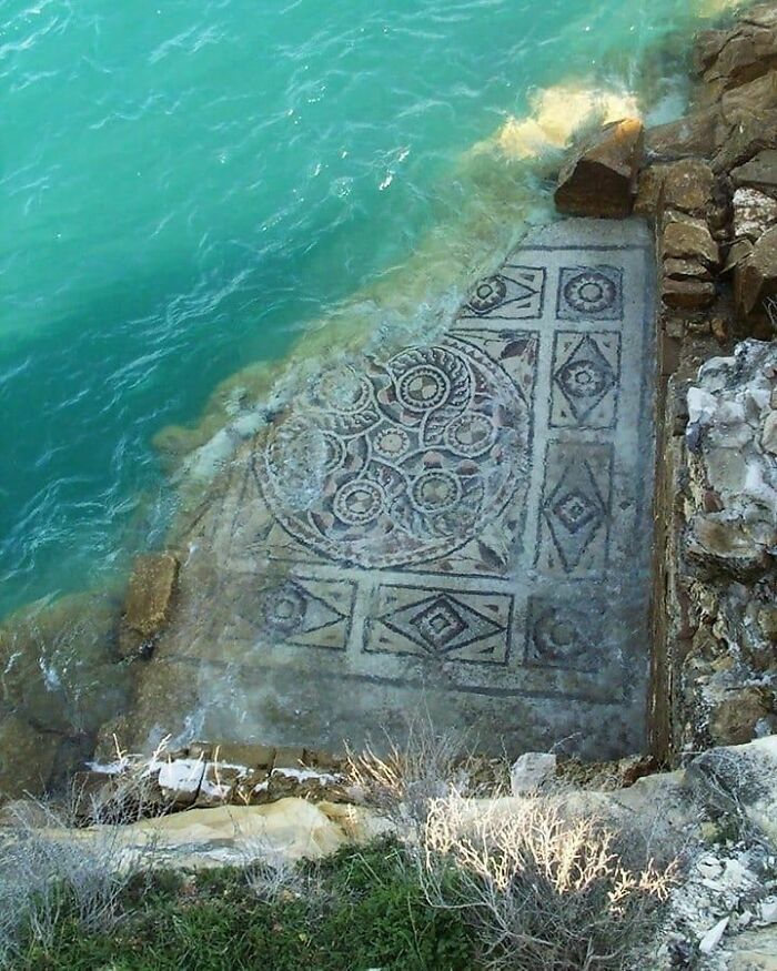 Ancient historical mosaic partially submerged in turquoise water near rocky shore