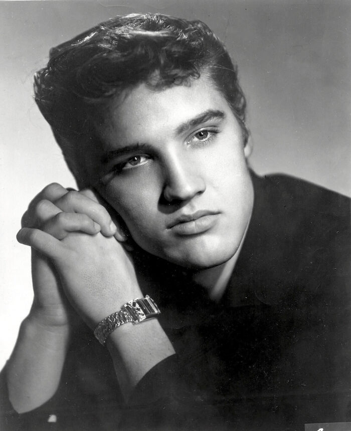 A young Elvis Presley in a black and white portrait, resting his chin on his clasped hands. Dive into useless facts.