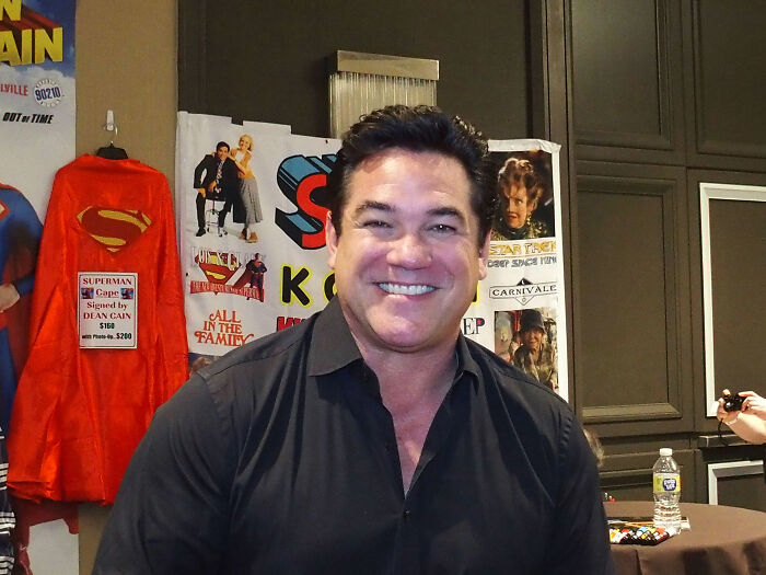 Actor Dean Cain smiling at event with Superman cape displayed behind