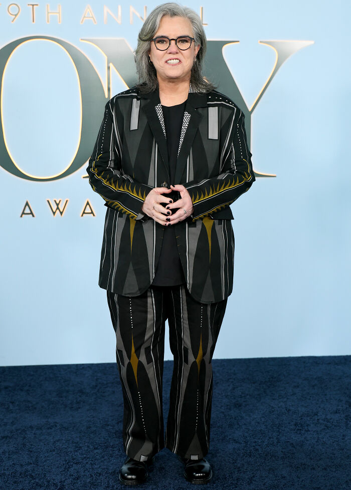 Person in black patterned suit and glasses posing on Tony Awards red carpet