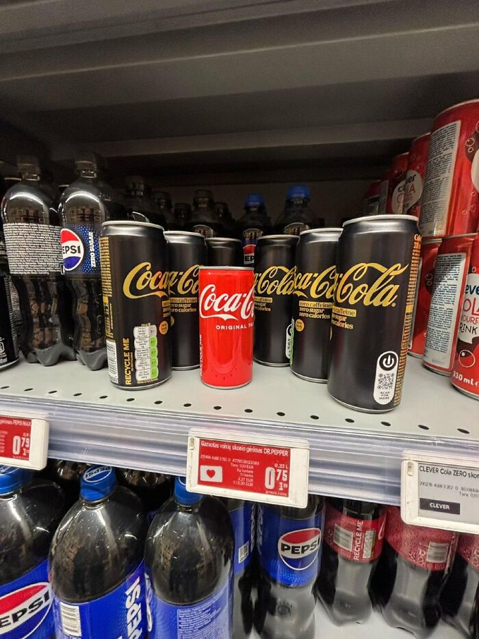 A single red Coca-Cola can stands out among several black Coca-Cola Zero Sugar cans on a supermarket shelf, a cringe image.