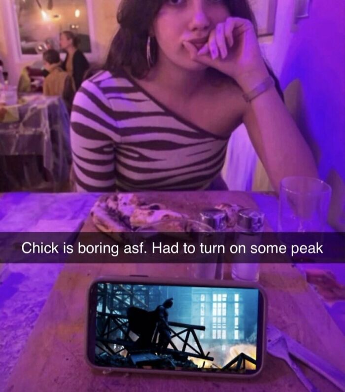 A woman at a restaurant with a phone showing a video, creating a cringe moment. The screen says Chick is boring asf.