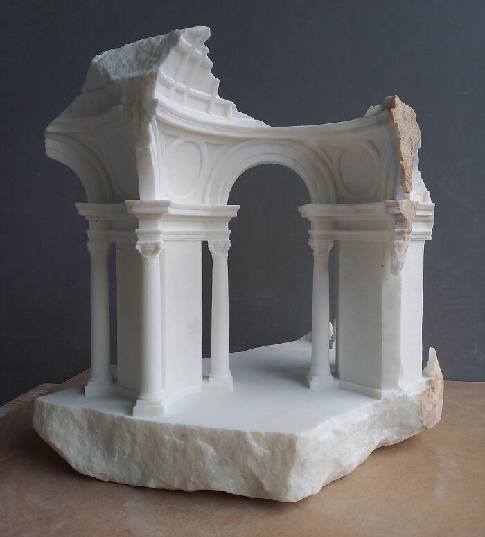 Artist carves tiny architectural models of ancient buildings into solid stone, displaying a grand archway with columns.