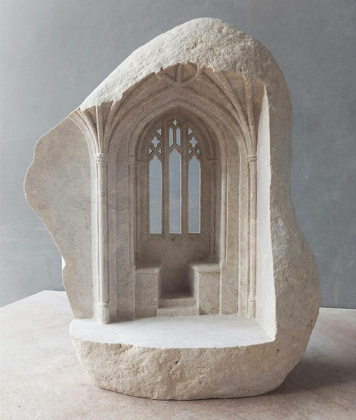 Artist carves tiny architectural model of a classical building in solid stone, featuring a grand window.