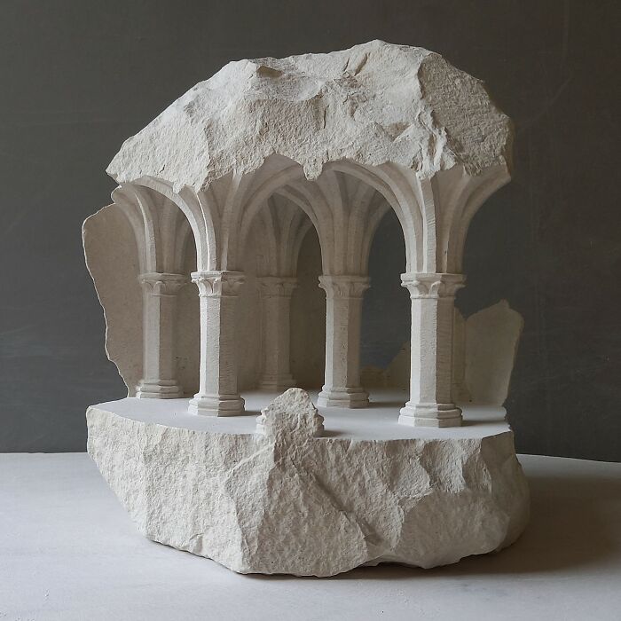 Artist carves tiny architectural models of classical buildings into solid stone, showing intricate arched columns within a raw stone.