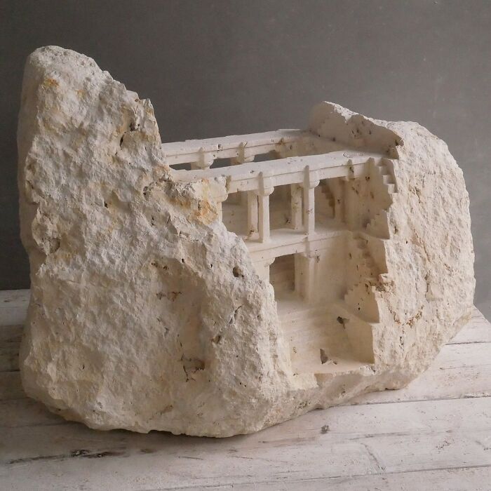 Artist carves tiny architectural models of classical buildings into solid stone, featuring a multi-level structure within a rock.