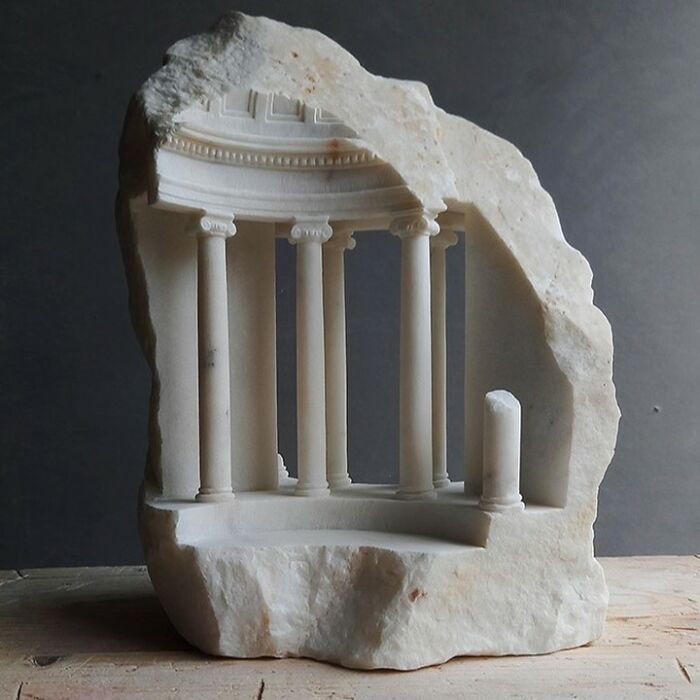 Artist carves a tiny architectural model of an ancient building into solid stone with a curved colonnade.