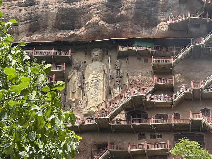 Travelers ascend wooden stairs alongside large Buddha carvings in a mountain cliff, highlighting travel in China.