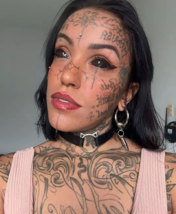 A woman with heavily tattooed face and chest, including her tattooed eyes, looks directly at the camera. She wears a black choker.