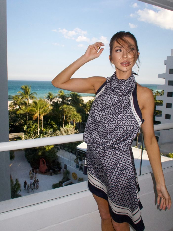 Miami Swim Week model in sleeveless printed dress poses on balcony with ocean and palm trees in background Miami Swim Week model in sleeveless printed dress poses on balcony with ocean and palm trees in background