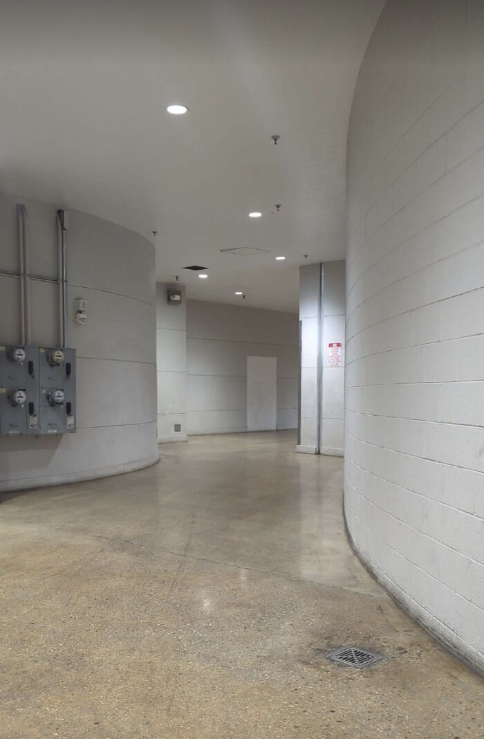 A curved, empty hallway with white walls and exposed pipes, evoking a haunting liminal space.