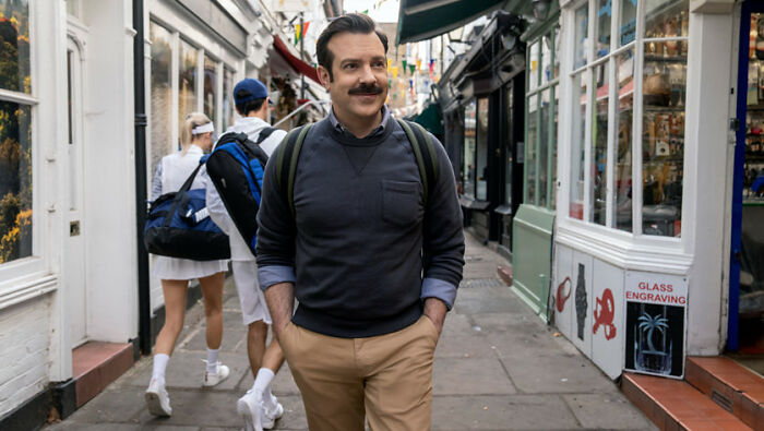 Jason Sudeikis walking down a street scene from Ted Lasso with a backpack and casual attire Jason Sudeikis walking down a street scene from Ted Lasso with a backpack and casual attire