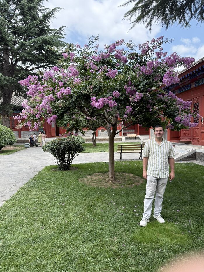 A traveler stands smiling beside a vibrant purple flowering tree in a traditional Chinese garden, showcasing China.