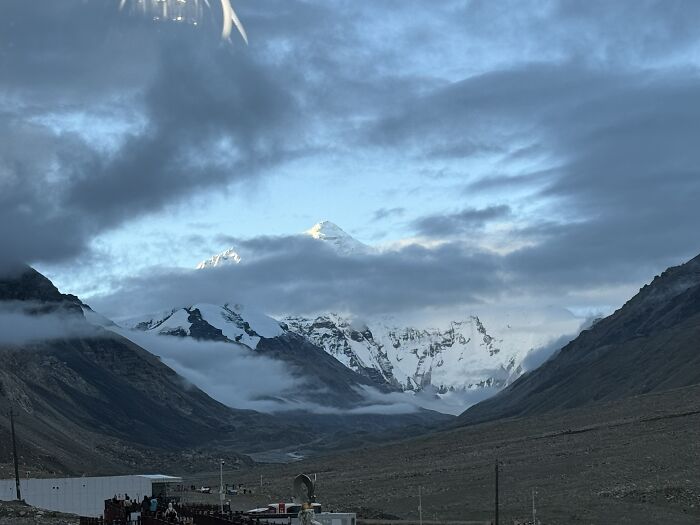 Majestic snow-capped mountains in China, partially obscured by clouds, with a valley and base camp structures below.