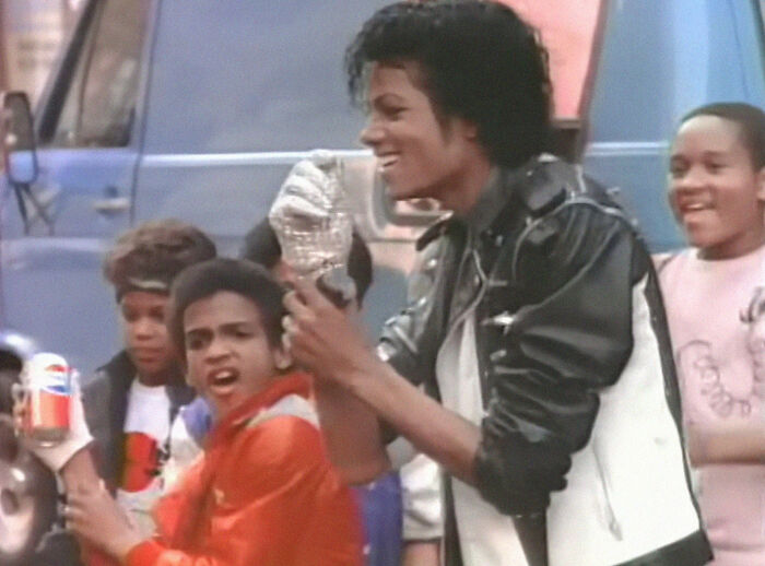 Michael Jackson wears a sequined glove and leather jacket, smiling next to kids, one holding a Pepsi can. Enjoy useless facts.