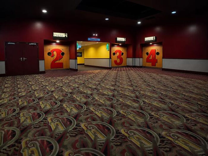 An empty movie theater lobby with red walls and patterned carpet, featuring numbered doors leading to cinema liminal spaces.
