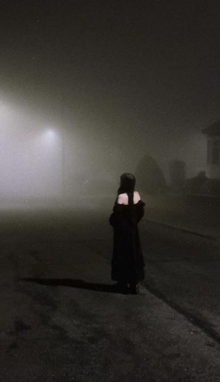 A lone figure in black stands on a foggy street at night, evoking a haunting liminal space.