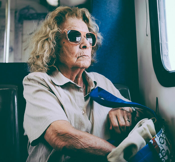 An 80-year-old woman with blonde curly hair and sunglasses, sitting on a public staircase with a bag, looking reflective.